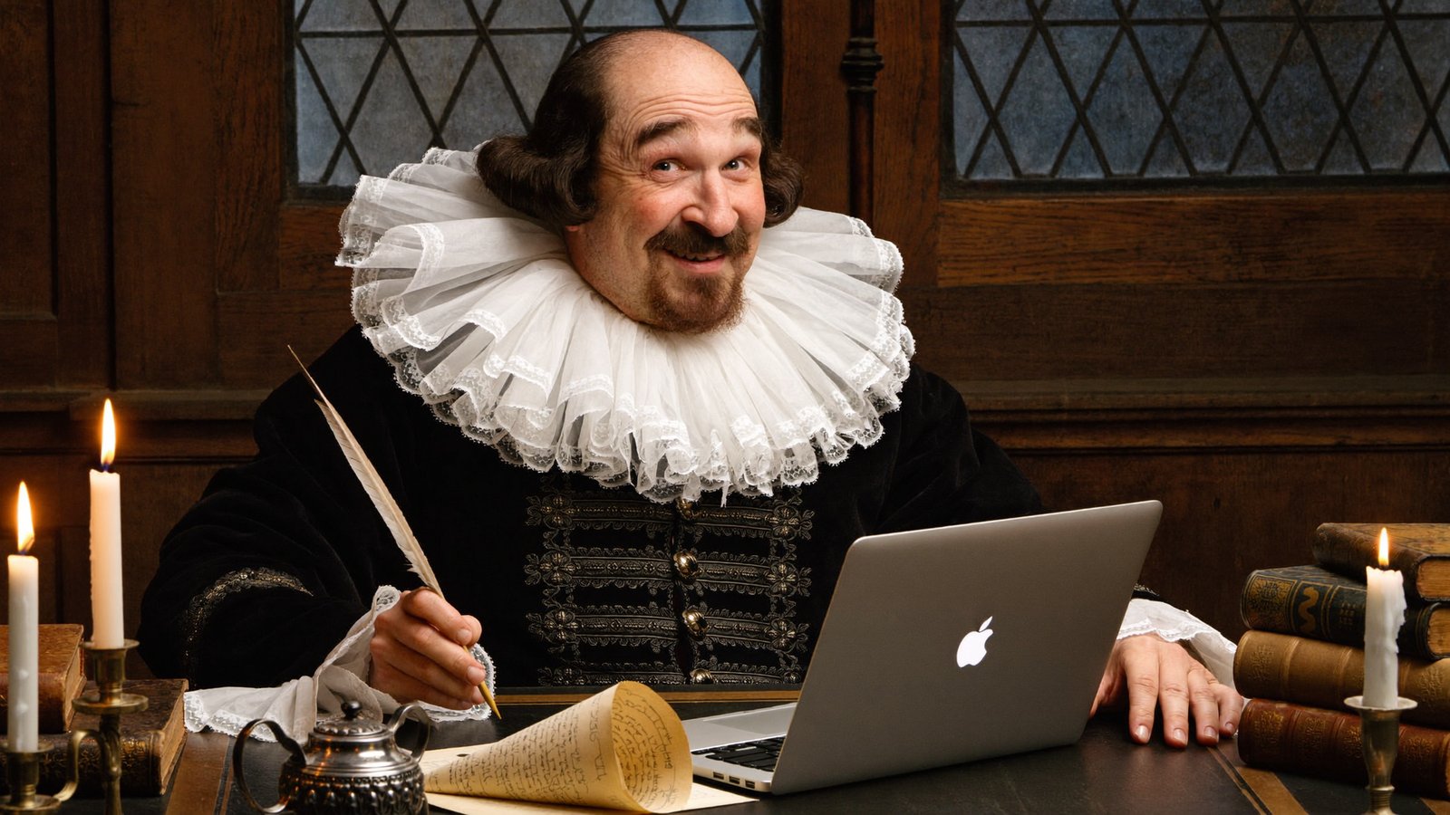 William Shakespeare at a candlelit Tudor desk, quill in hand, writing on a MacBook and grinning at the camera